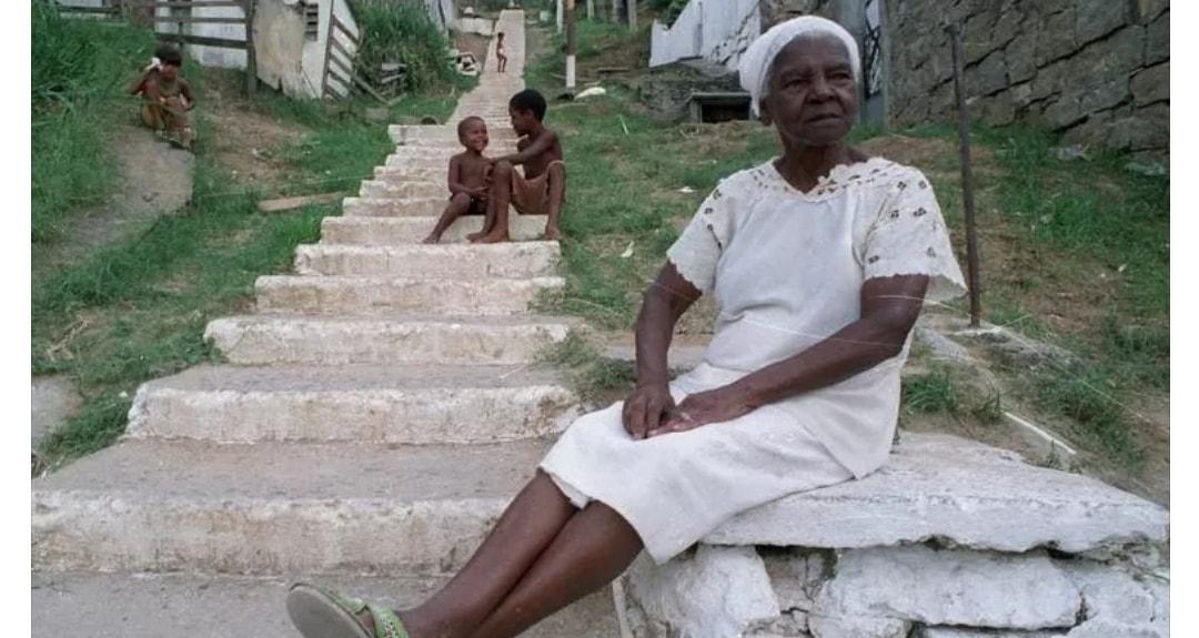 Em primeiro plano há a imagem de uma mulher negra, idosa,  sentada com os braços sobre as pernas, com vestido e lenço na cabeça brancos. Em segundo plano, há uma escadaria onde encontram-se sentados dois meninos negros conversando.
