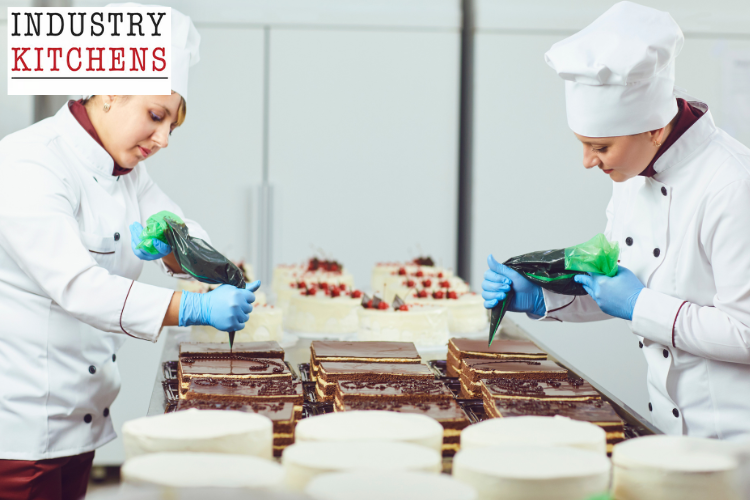 Professional pastry chefs in white uniforms and chef hats decorating cakes with chocolate in a commercial kitchen.