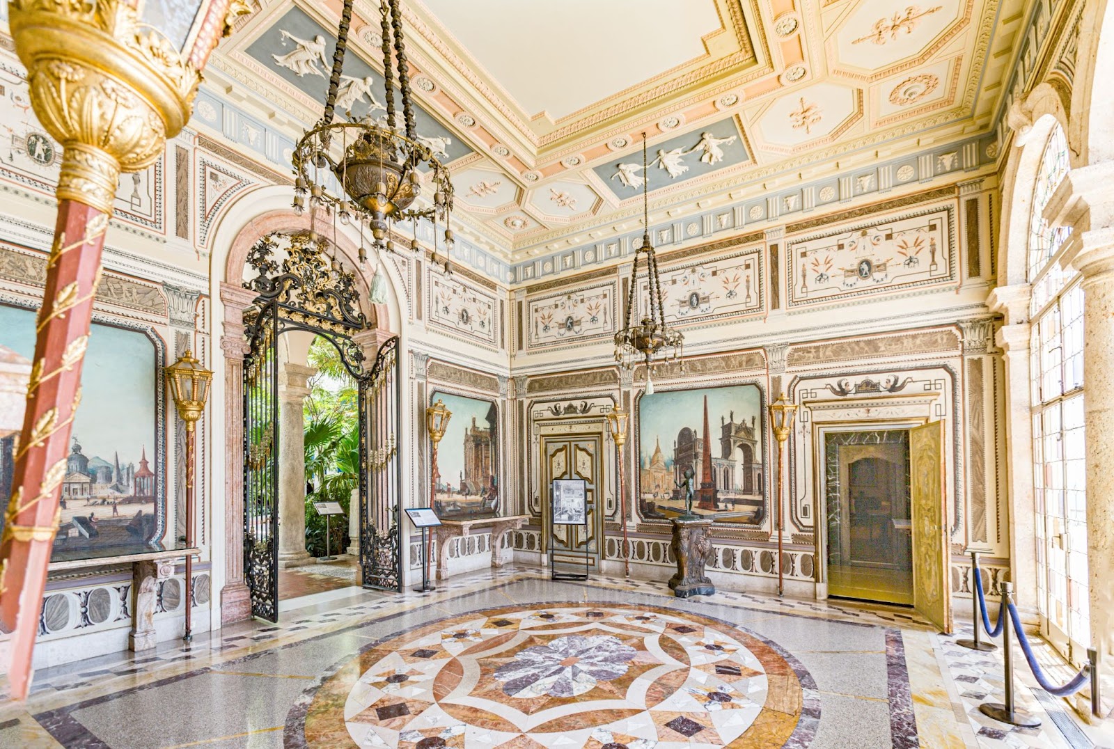 Interior view of the Vizcaya Museum and Gardens in Miami featuring elaborate neoclassical decor, frescoed walls, hanging chandeliers and a marble mosaic floor with a central medallion design.