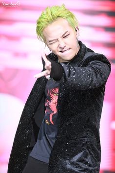 This contains an image of G-dragon   with green hair and black jacket pointing to the side while standing in front of a pink background