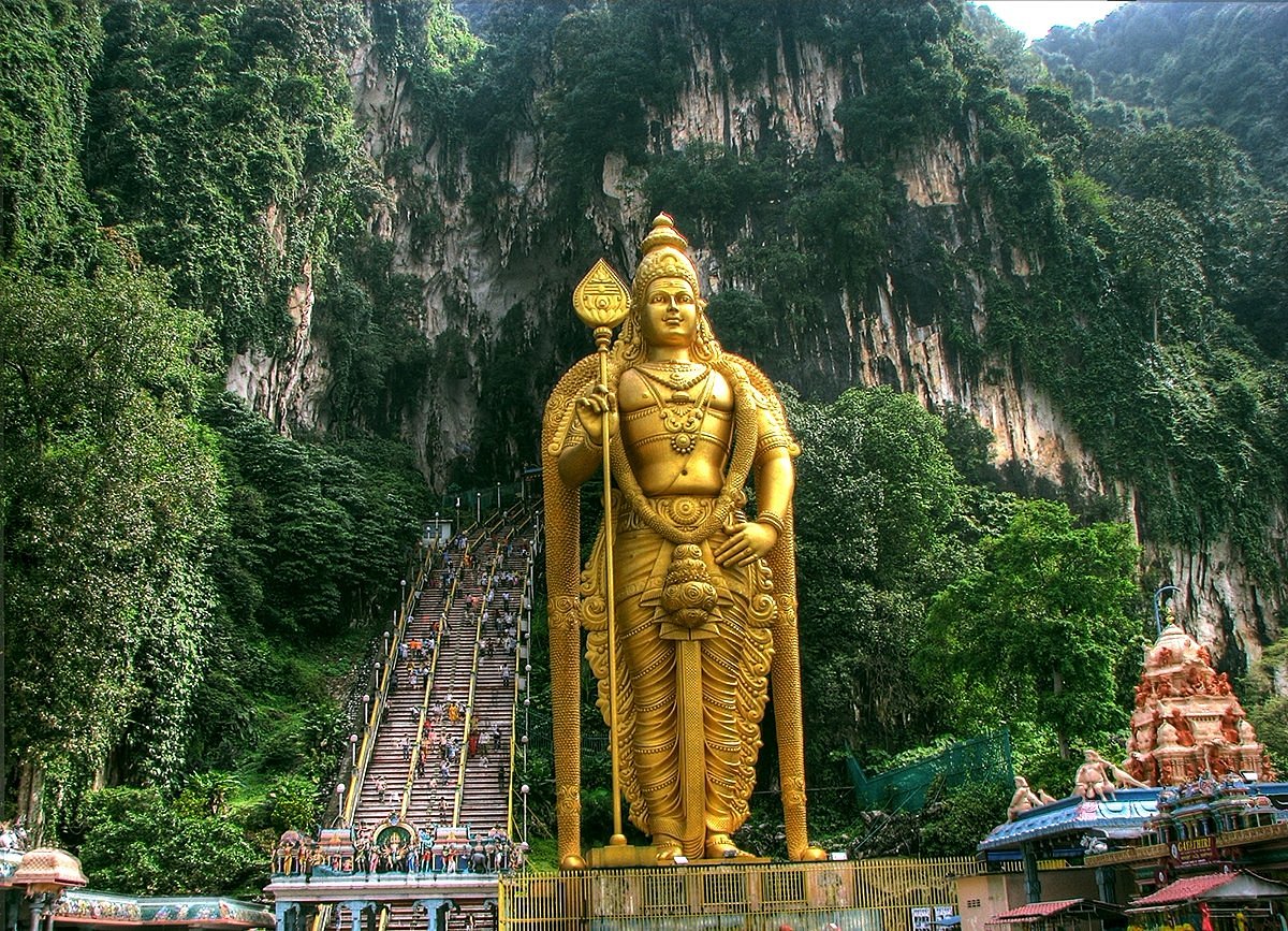 The iconic golden statue of Lord Murugan at the entrance of Batu Caves in Malaysia, surrounded by lush green limestone cliffs and a staircase filled with visitors.