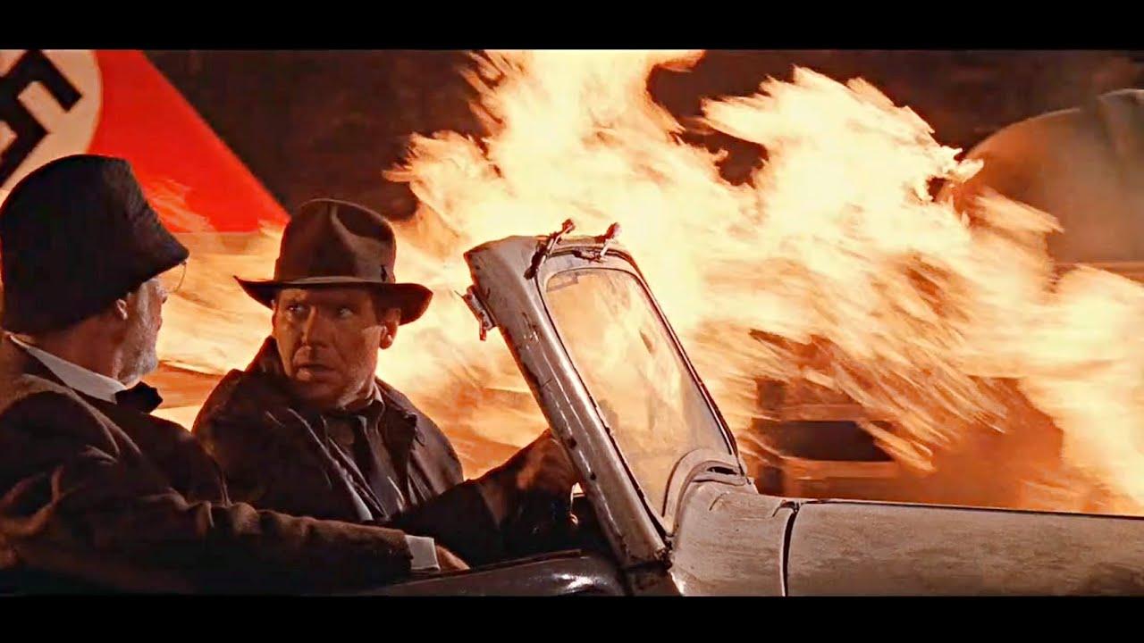Indiana Jones does a befuddled take to his dad while driving a car next to the plane engulfed in flames.