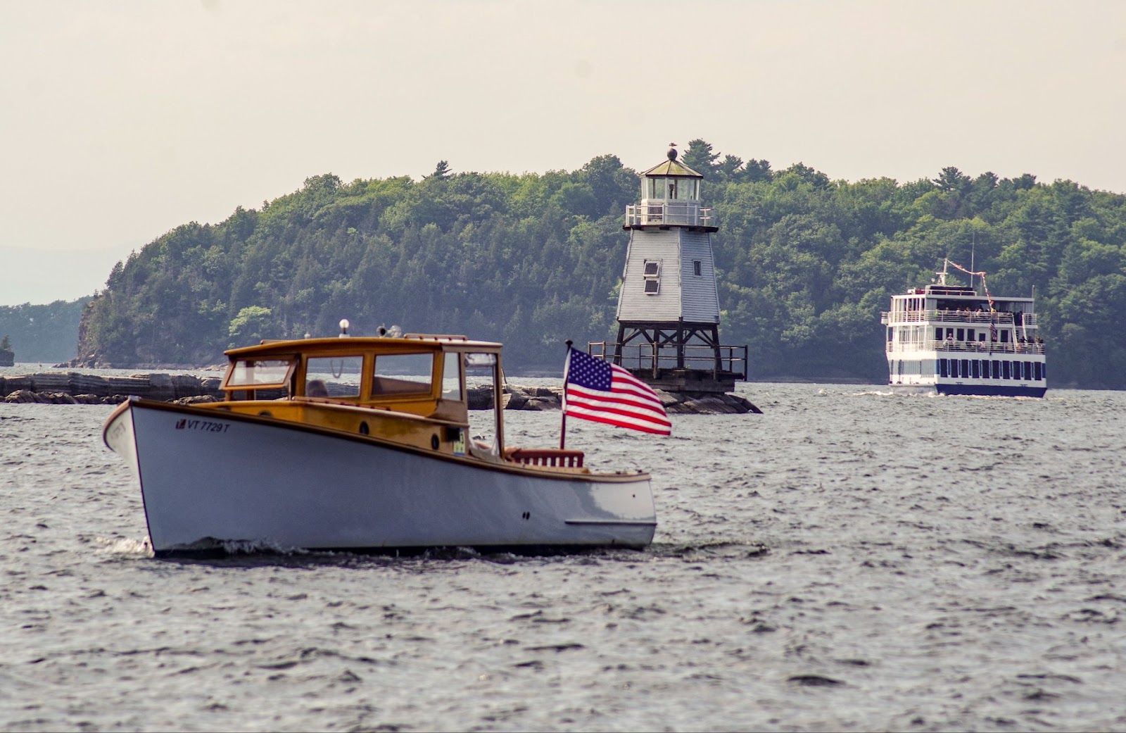 Summer on Lake Champlain with an American flag on an antique boat with Spirit of Ethan Allen in the background