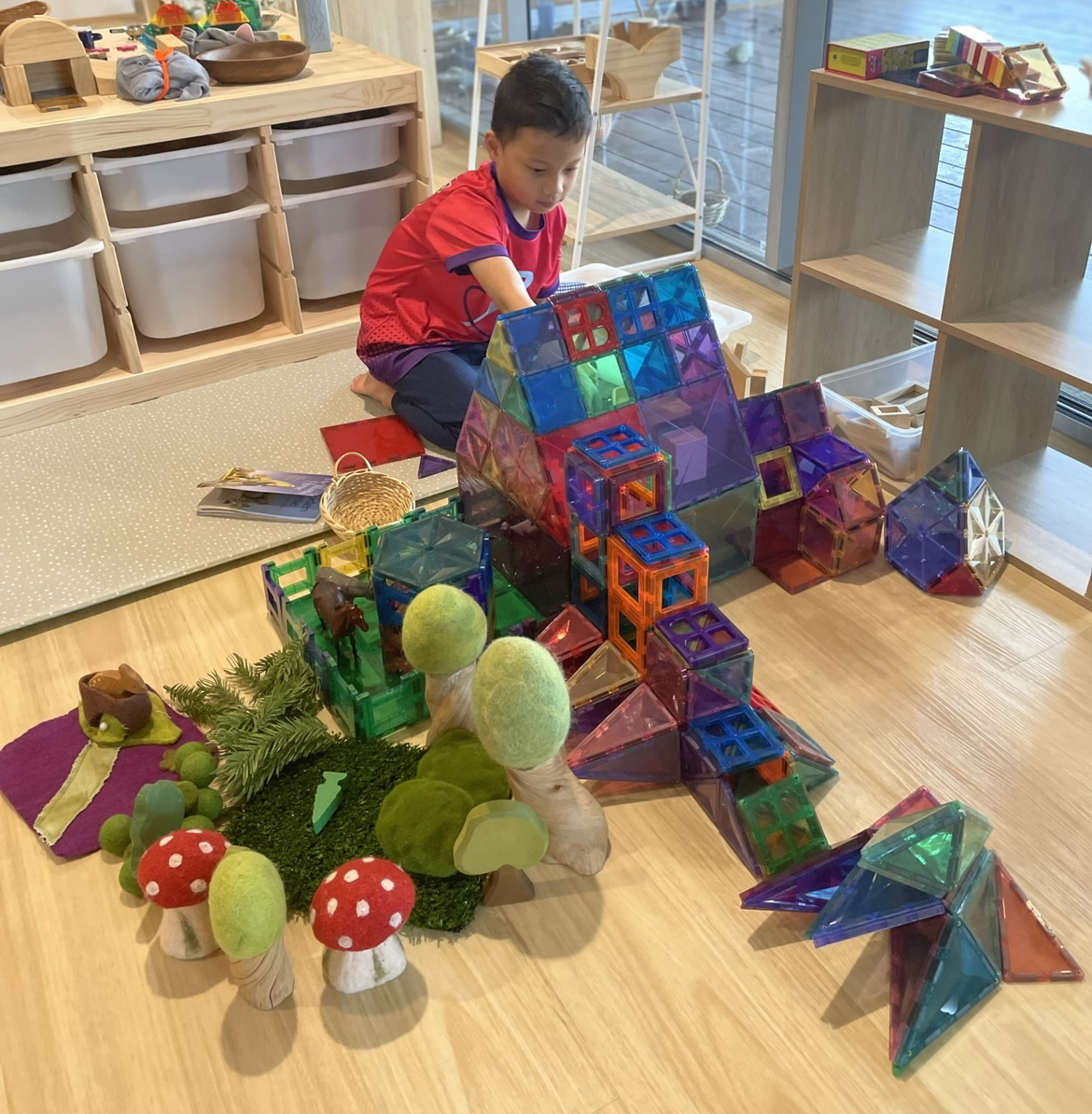 An older boy is building an elaborate structure with magnetic tiles, incorporating imaginative elements like trees, mushrooms, and a farm area. Alongside the grounded creations, he has also constructed a flying structure, blending creativity and storytelling in his playful scene