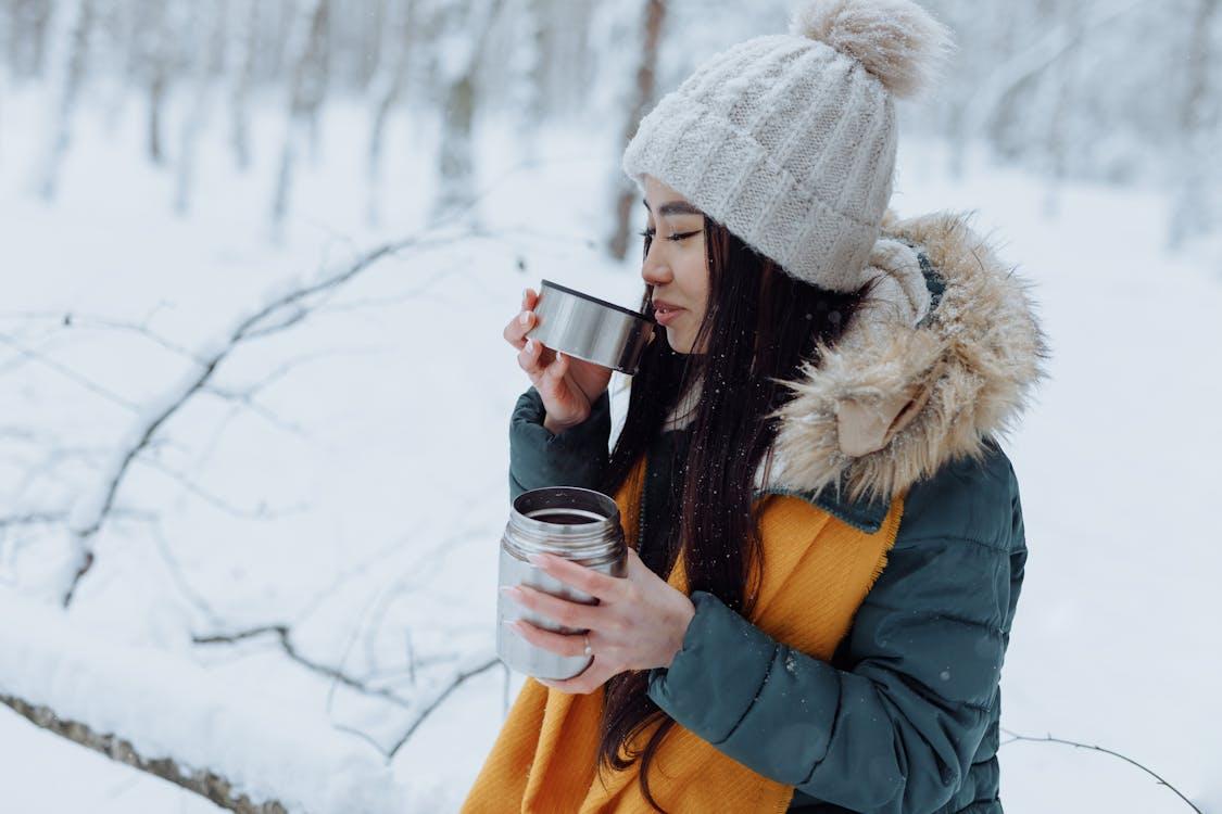 Free Asian woman sipping from a thermos in a snowy winter forest, staying warm and cozy. Stock Photo