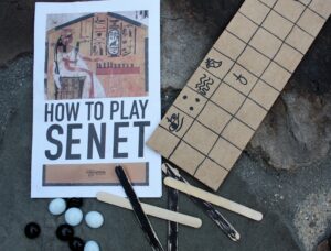 An instruction booklet labeled "How to Play Senet" and a wooden board decorated with hand-drawn hieroglyphs. Black and white round playing pieces and black and plain craft sticks are scattered around. 