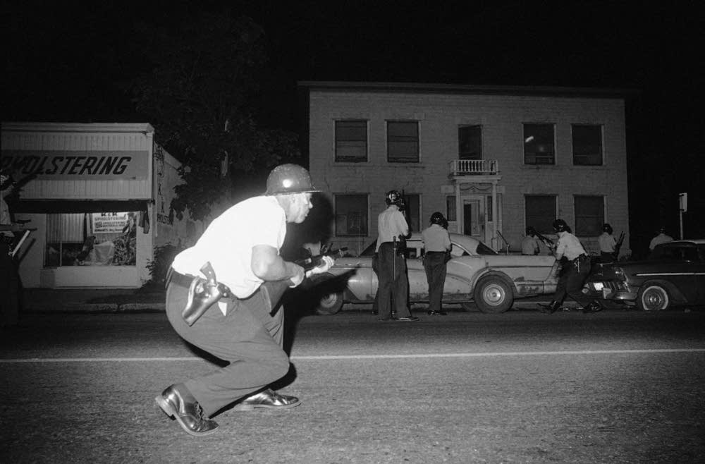 Image: Minneapolis Police during the Plymouth Ave Riots, 1967. Photo by Robert Walsh, circa 1967. A black-and-white photo showing a policeman crouching in the foreground while holding a gun. In the background are several policeman standing around cars on a city street.