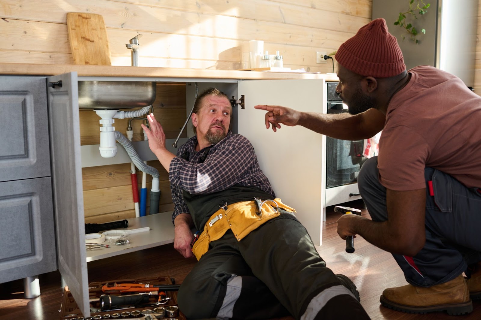 A senior plumber guides his young colleague while inspecting a sink pipe, discussing plumbing prices and cost-effective repair solutions.