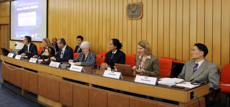 A panel of nine people, both men and women, dressed in formal attire, sit at a long wooden desk in a conference room with wood-paneled walls and a presentation screen in the background.