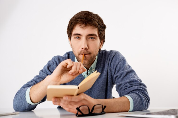 Thoughtful, creative male journalist, biting a pencil and holding a notebook