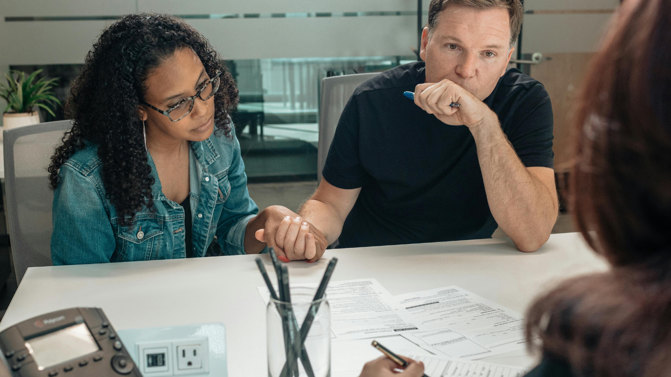 Serious couple holding hands while discussing legal documents with a professional during a formal meeting in an office setting.