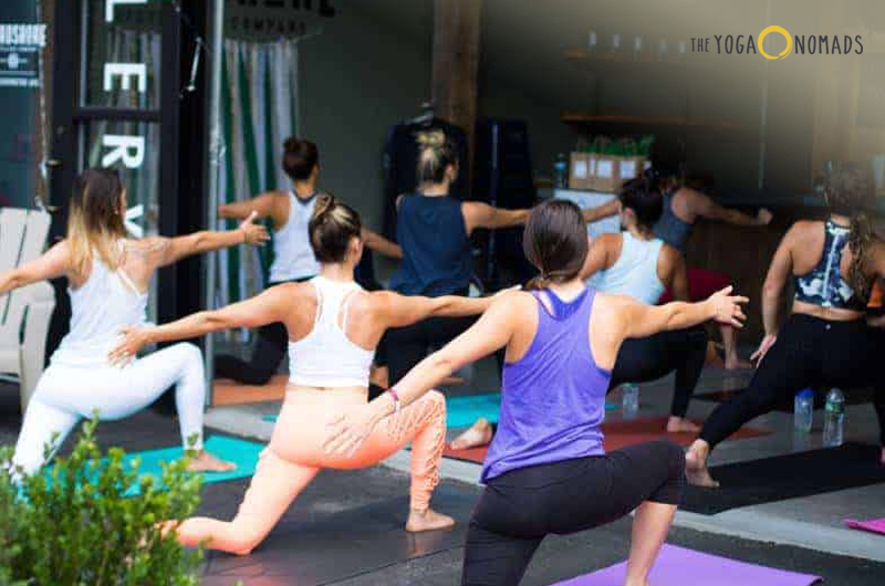 A group of individuals participating in a yoga class. They are in various yoga poses on mats, indicating the setting is likely a fitness or wellness center. The focus and alignment of the participants suggest it’s an organized group activity.