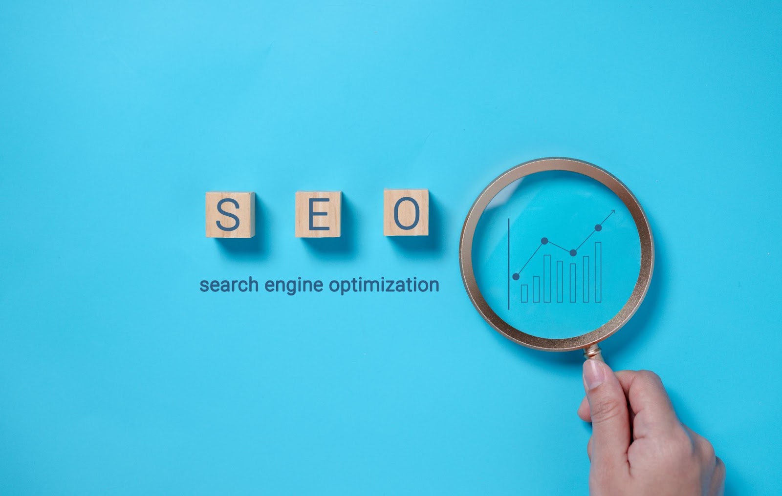 Word “SEO” written on wooden blocks over the words “search engine optimization” and a hand holding a magnifying glass over a graph. 