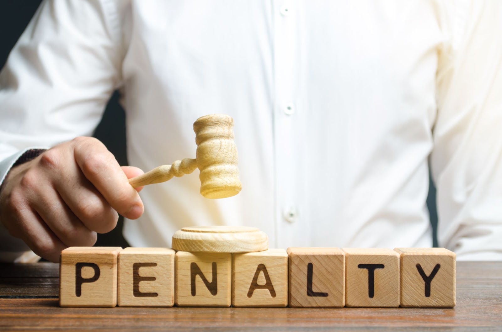 A cropped shot of a judge in a white long-sleeved polo holding a gavel above blocks that spell out the word "penalty," emphasizing the obligation to pay a fine or penalty.