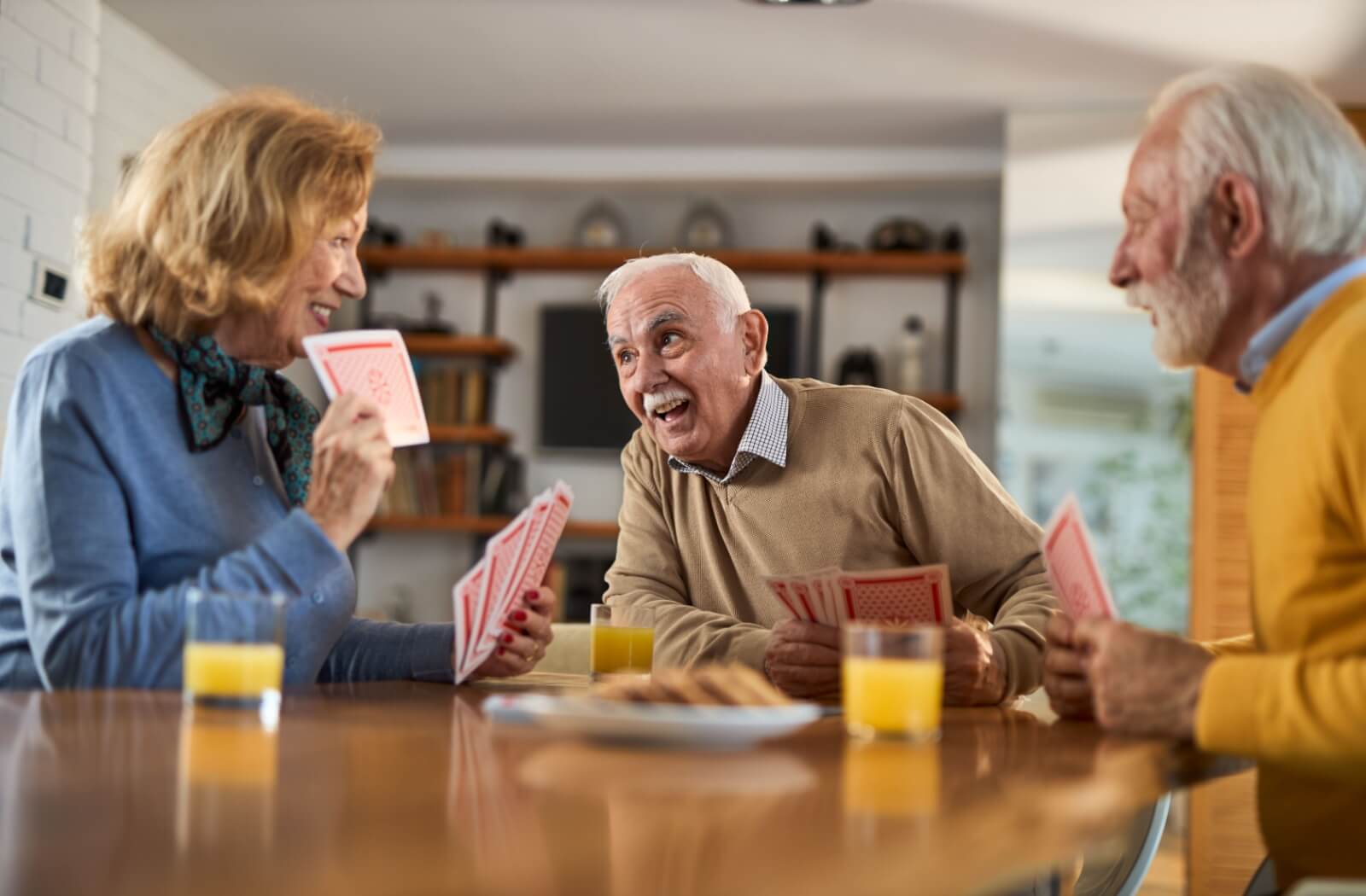 A group of happy seniors play cards and share a snack in the communal area of their senior living community.
