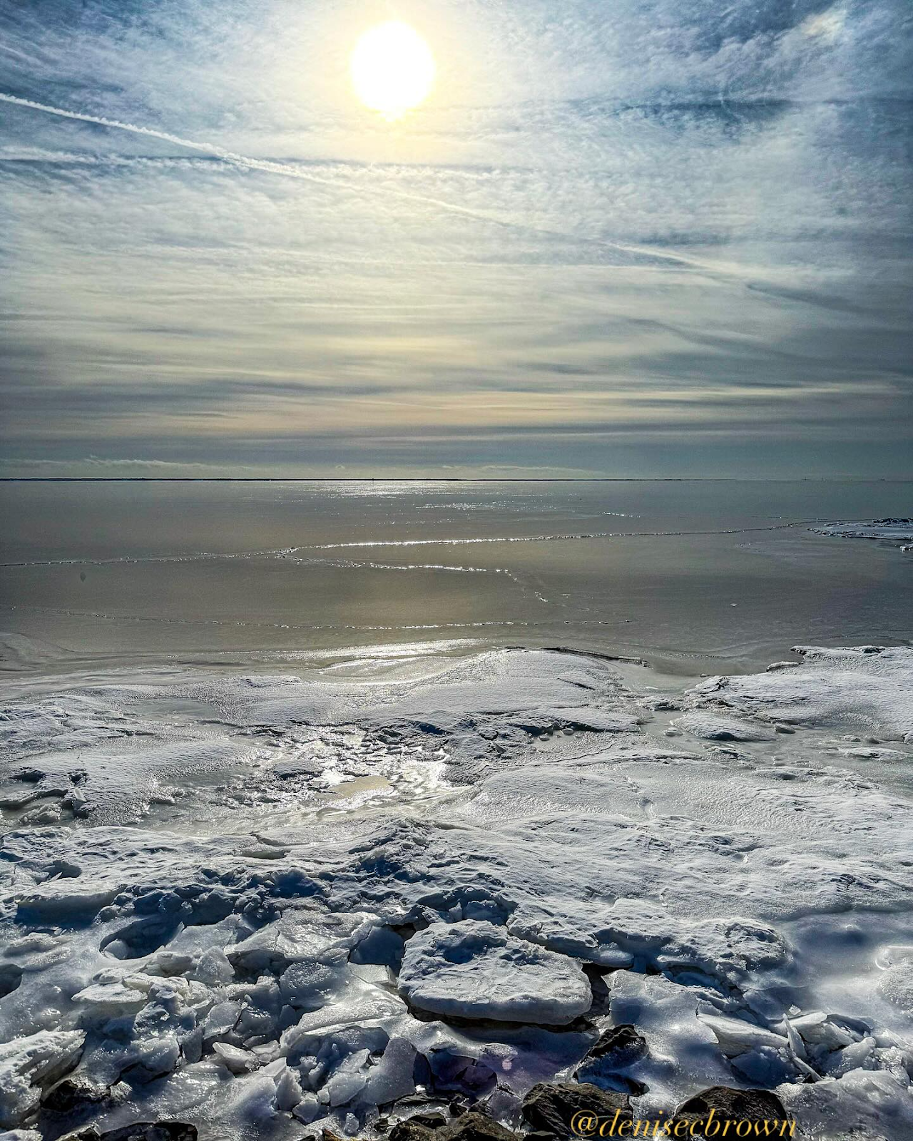 A serene winter landscape featuring a bright sun above icy terrain, with a mix of clouds and light reflecting off the surface. Captured by @ceramictilegirl1.