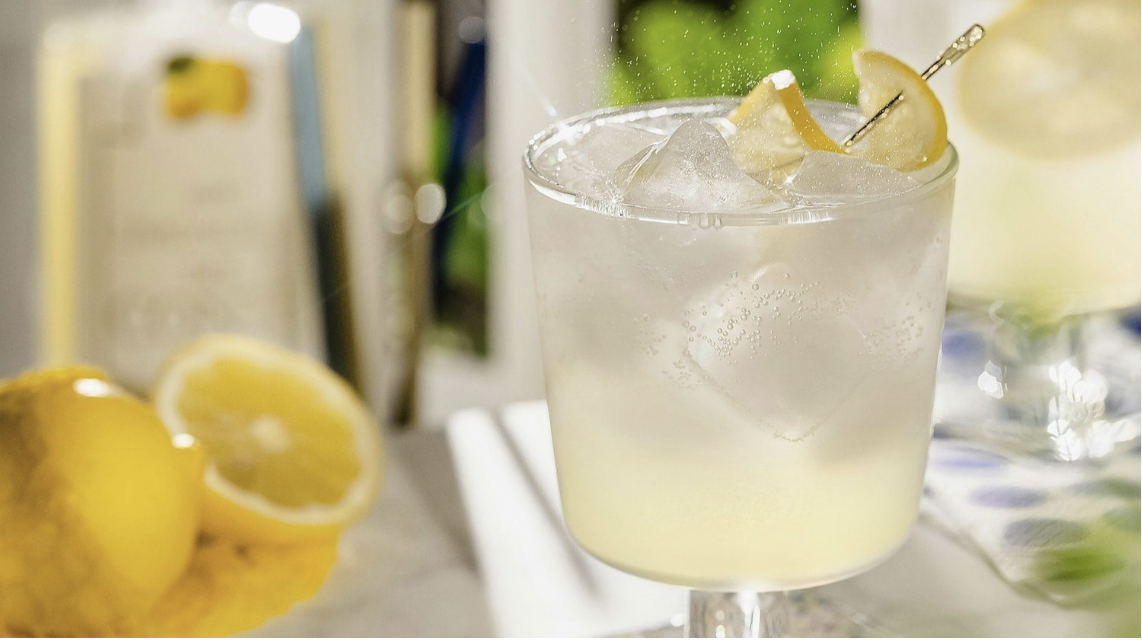 A limoncello spritz fills a glass with ice and a lemon garnish with lemons and a glass bottle in the background