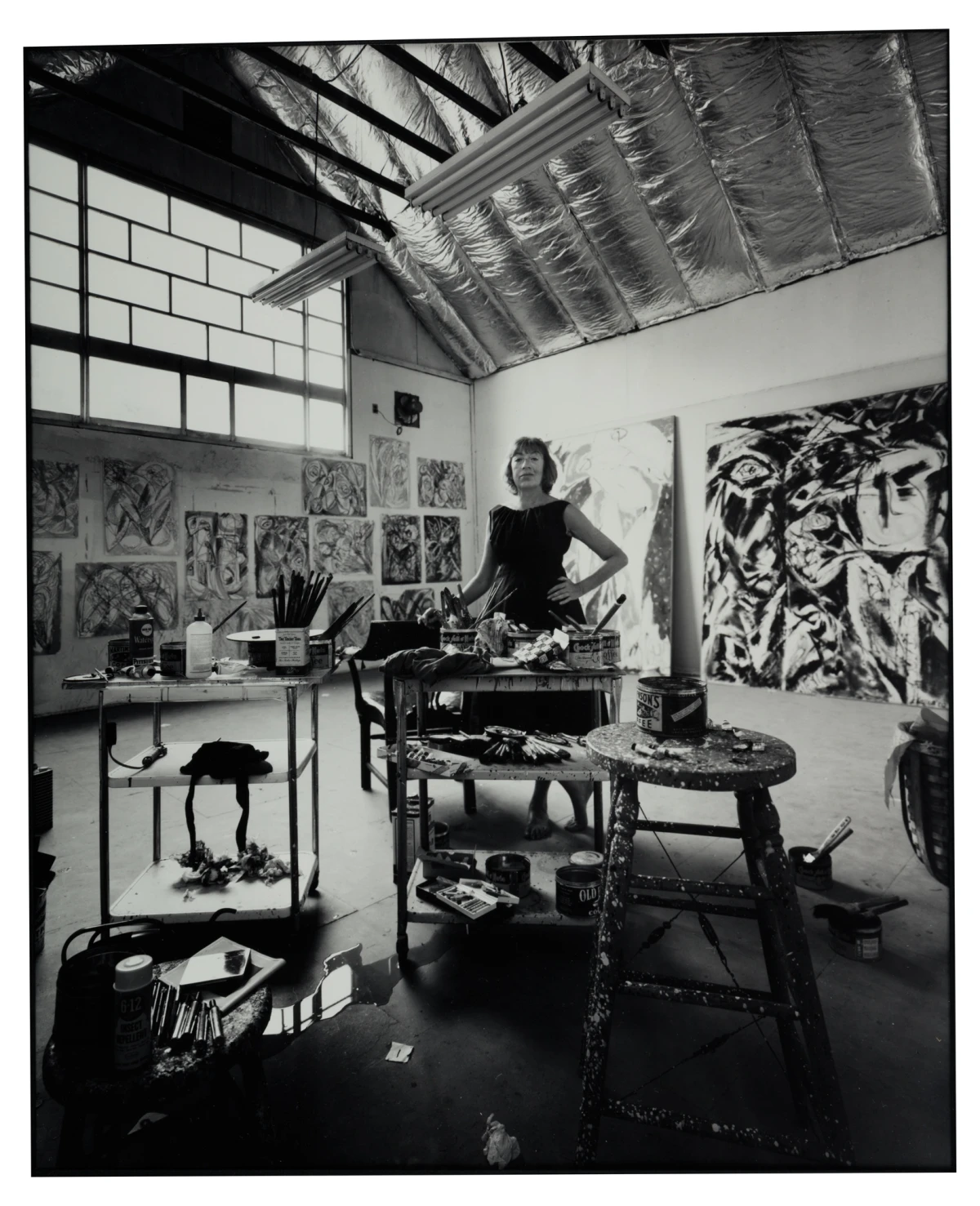 Lee Krasner standing in her art studio surrounded by abstract expressionist paintings, paint cans, brushes, and art supplies, with large canvases and natural light highlighting her creative workspace, reflecting her role as a pioneering female abstract artist in postwar American art.