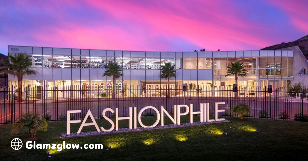 A modern, glass-fronted building with large windows and a sleek design, illuminated under a vibrant pink and purple sunset. The building is labeled 'FASHIONPHILE,' with the brand name prominently displayed on a large sign in front of the structure. Palm trees and well-manicured landscaping surround the building, with the logo 'Glamzglow.com' in the corner.