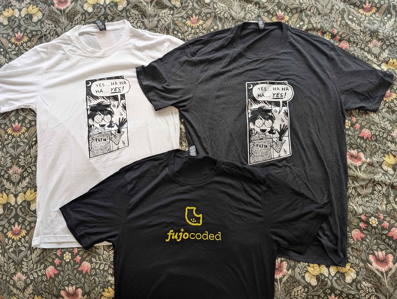 Three tee shirts laid flat. The white and grey shirts have the “Yes…Ha Ha Ha…Yes!” design with Boba-tan pressing her face to a window, while the black shirt has a bright yellow FujoCoded logo.