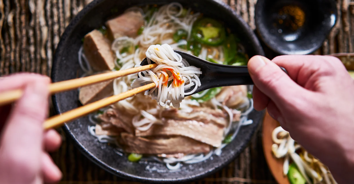 (Close-up of hands using chopsticks and a spoon to lift rice noodles from a bowl of Vietnamese pho with beef slices, green chili, and broth.)