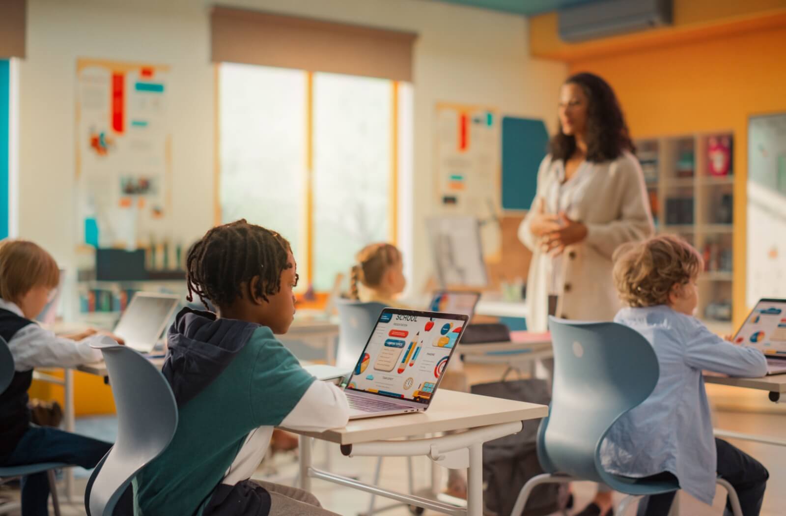 A bright classroom full of kids using laptops as their teacher presents a lesson.