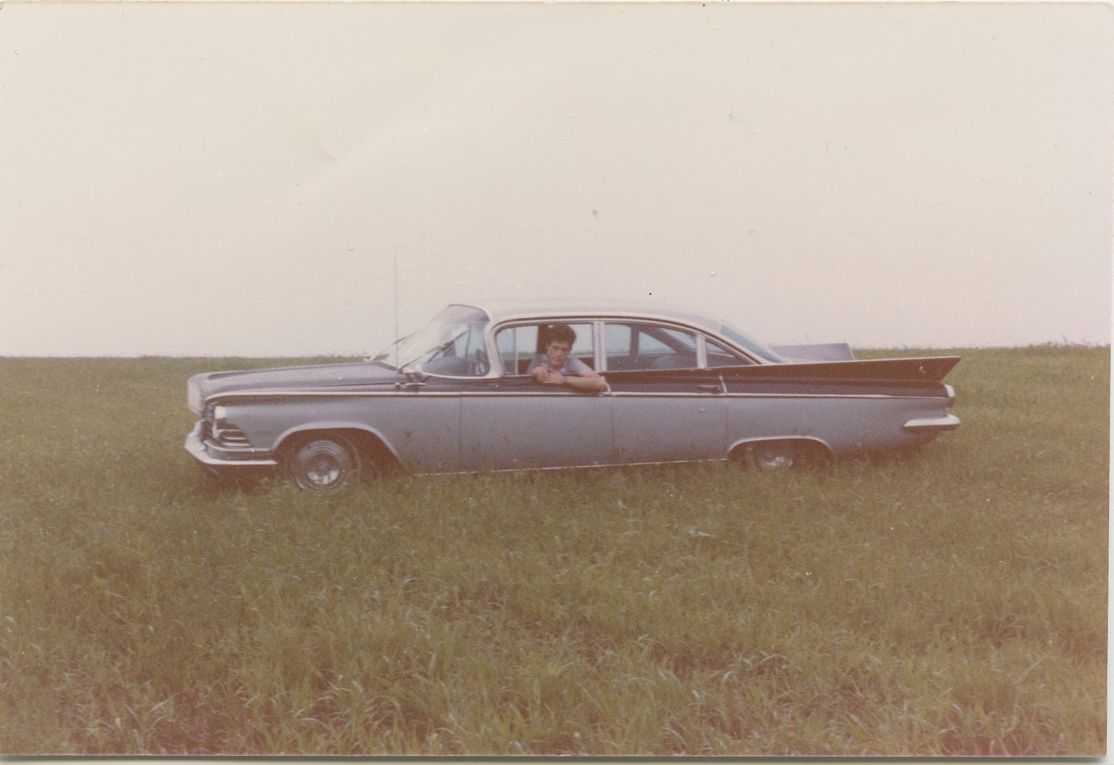 A blue car in a field, with a man leaning slightly out of the driver's seat window