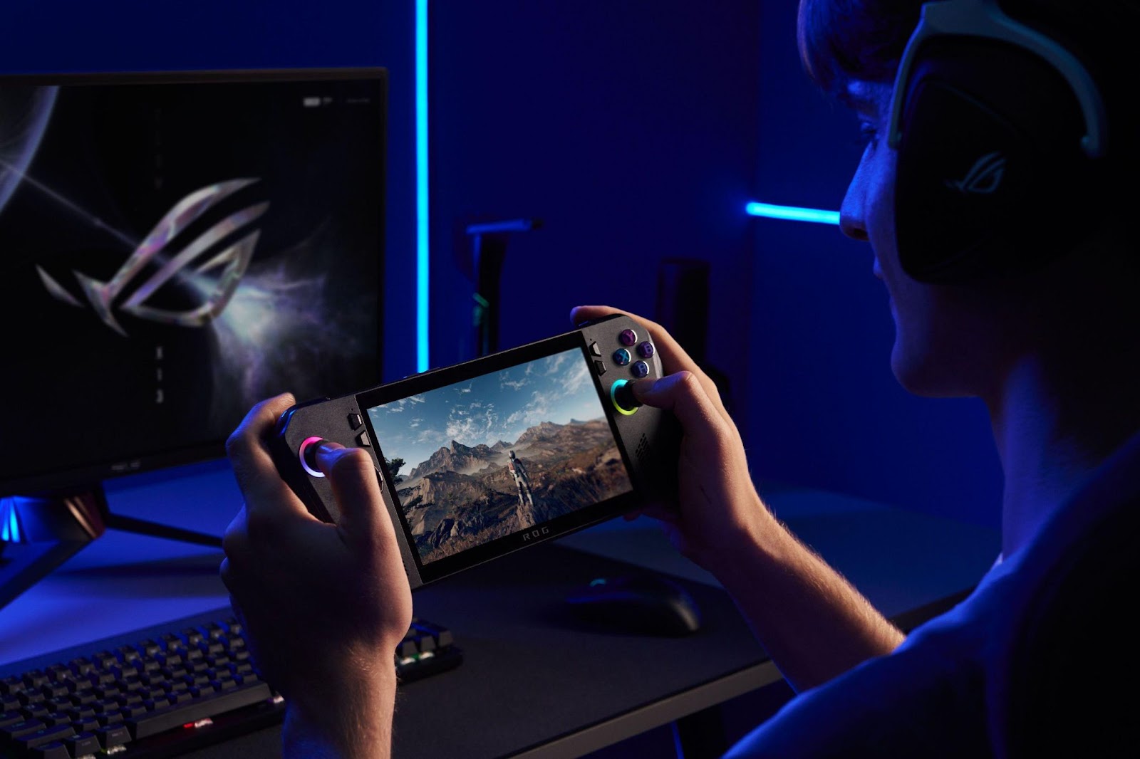 Gamer segurando rog ally x, teclado mecânico iluminado e monitor ROG em ambiente neon azul.