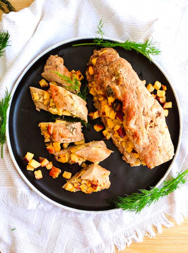 Sliced pork tenderloin stuffed with apples and herbs, served on a black plate with fresh dill.