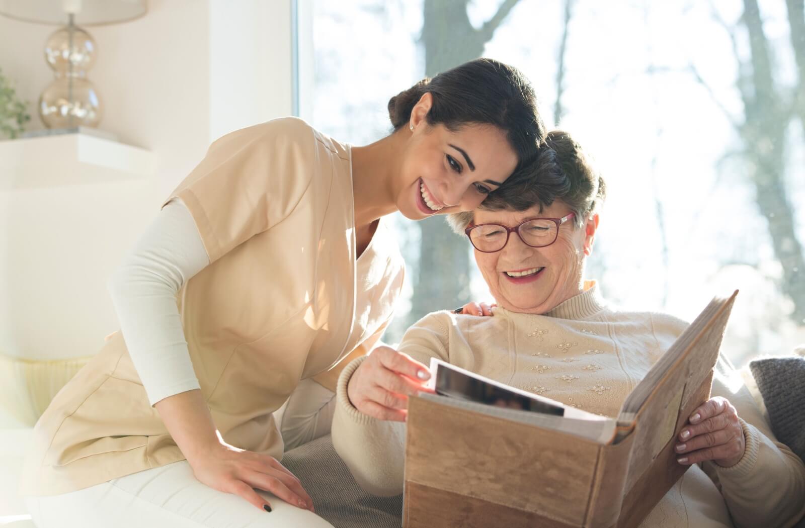 A smiling caregiver looks through a senior's photo album with them and asks engaging questions about what they're looking at.