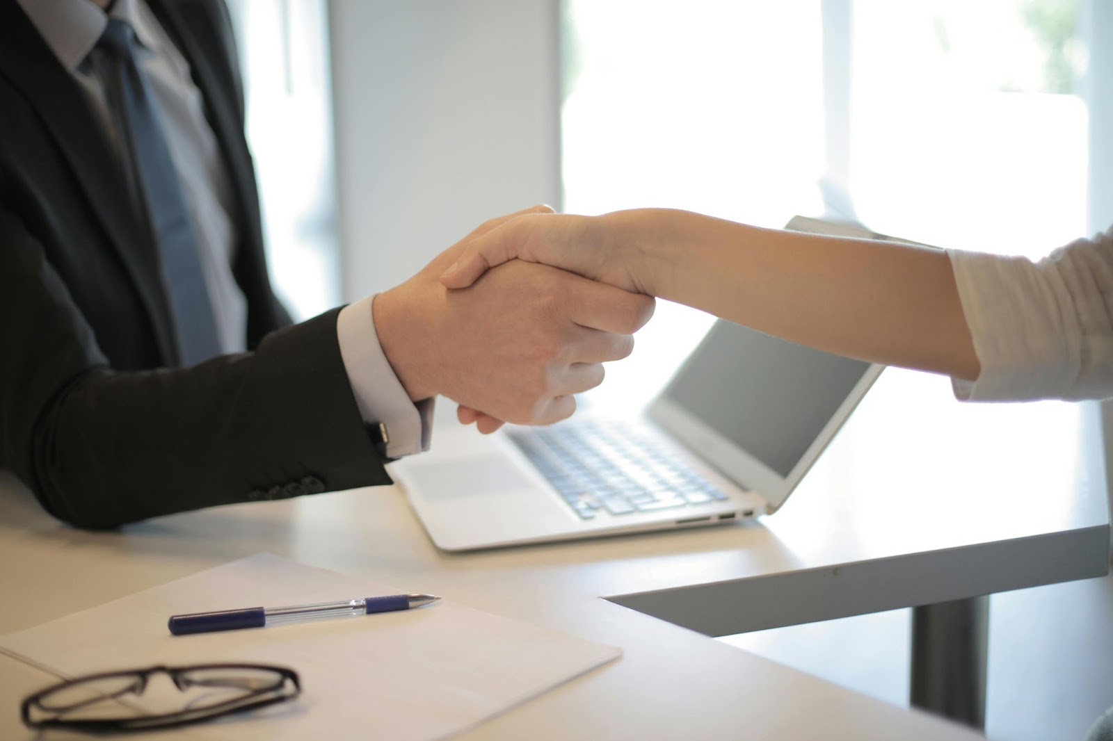 Two white people in business clothes shake hands in a brightly lit office