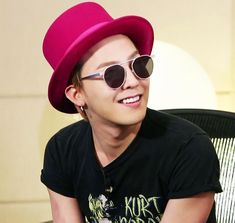 This contains an image of G-dragon wearing a black t-shirt and a red cap with glasses