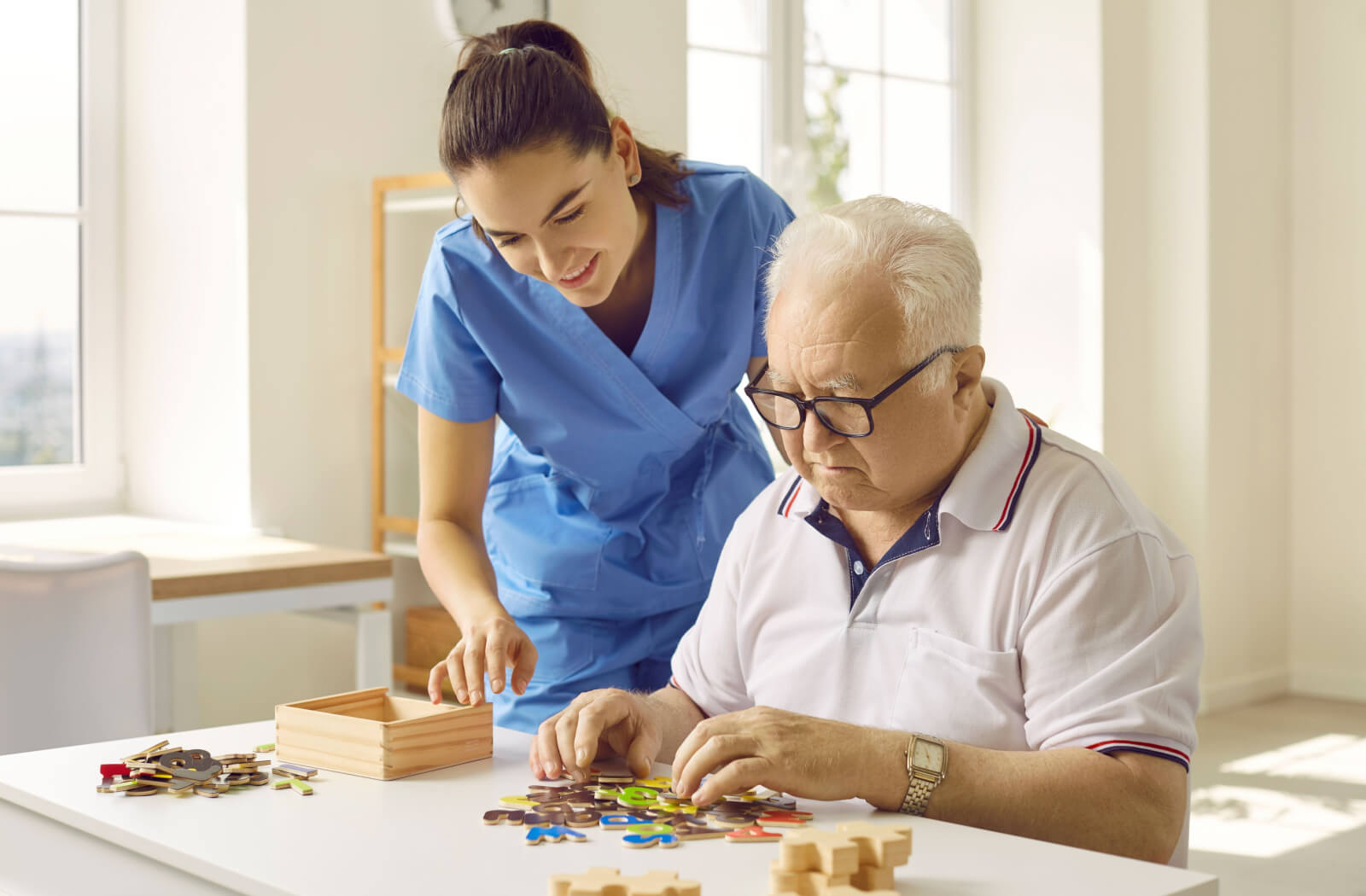 A caregiver smiling while standing beside a resident with Lewy body dementia in memory care while they work together on a puzzle.