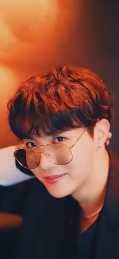 This contain BTS J-Hope with red hair and glasses posing for a photo in front of an orange background