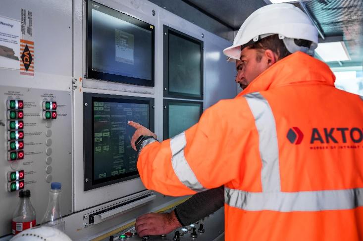 A person in an orange jacket and white hardhat pointing at a screen  Description automatically generated