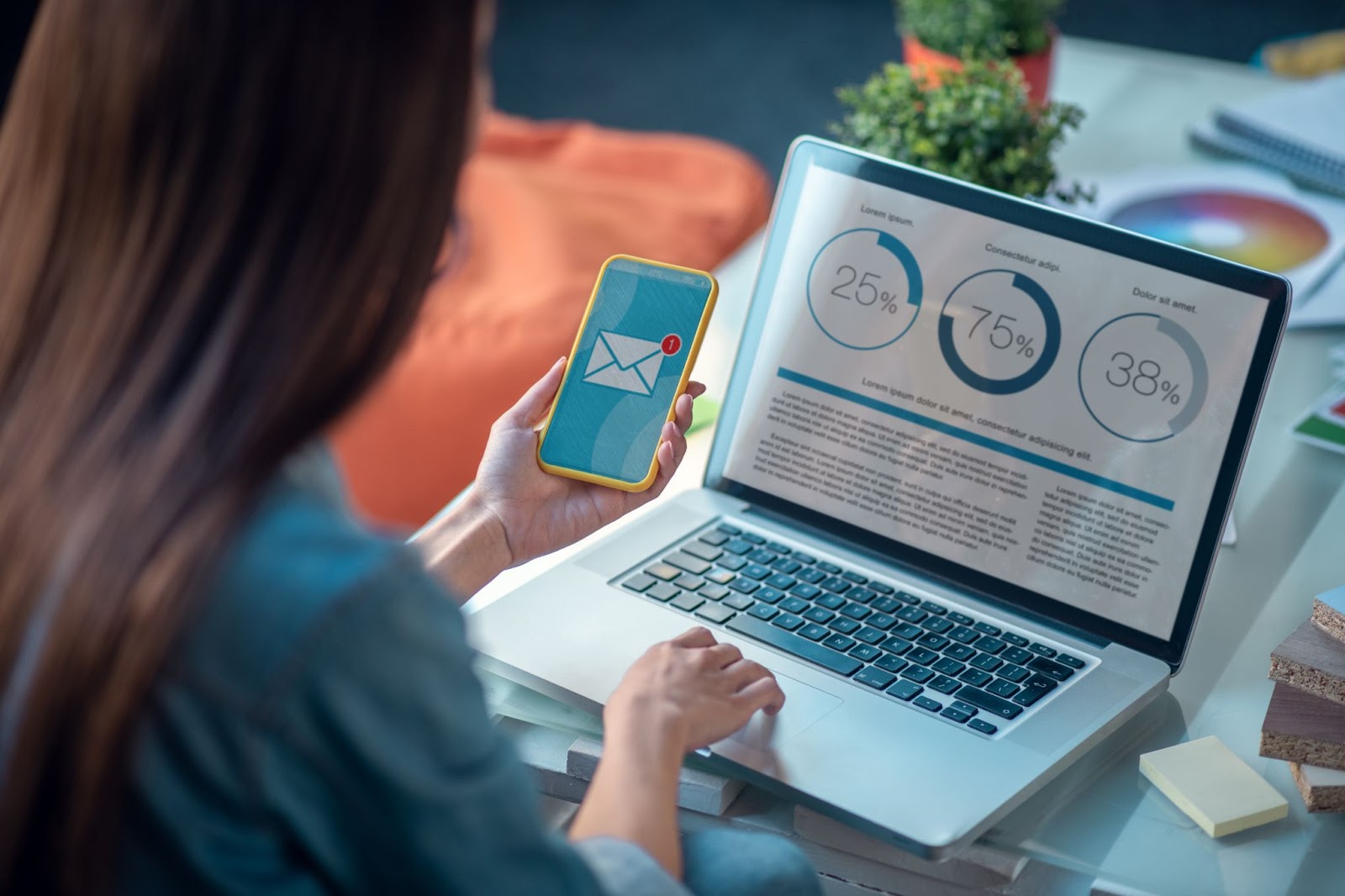 Woman holding a phone with an email icon and looking at data on a laptop. 