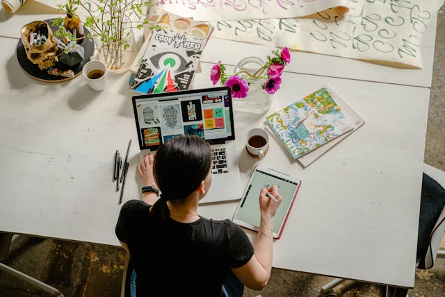 A woman focused on her laptop, sitting at a desk surrounded by books and a cozy workspace vibe.