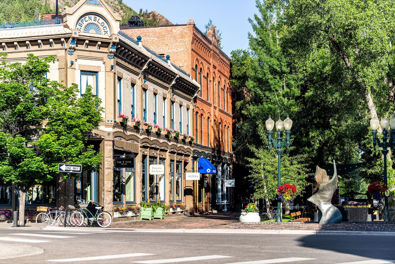 Street view of downtown Aspen, Colorado, showing historic brick buildings, boutique storefronts, bicycles and lush green trees on a sunny day.