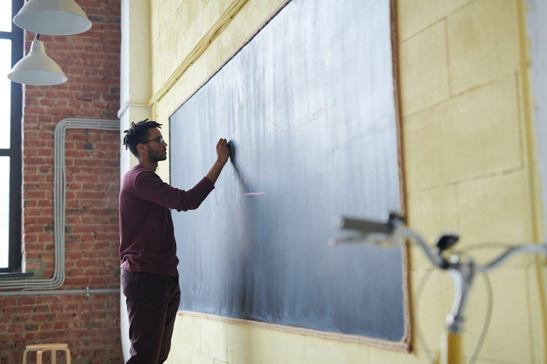 Free Man with dreadlocks writing on a blackboard in a classroom setting. Stock Photo