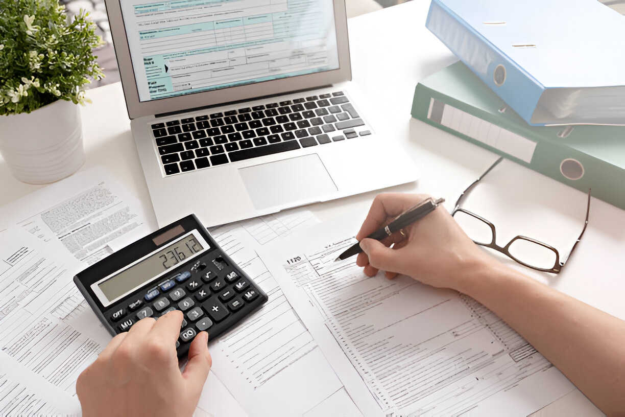 Person filling out tax forms with a calculator and laptop, preparing financial documents for tax filing.