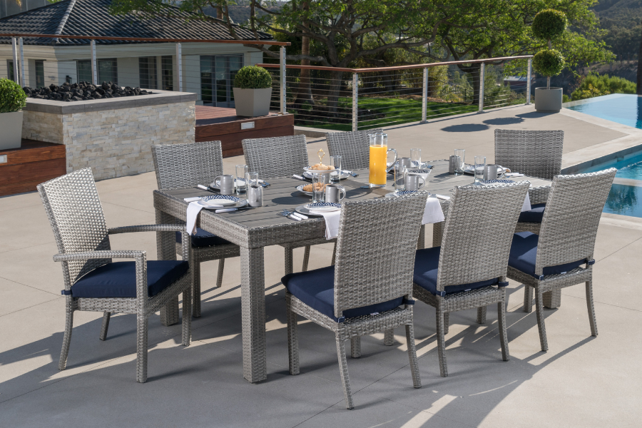 A cozy outdoor seating area with navy blue Sunbrella® cushions on wicker chairs and a sofa, centered around a rectangular fire table with vibrant flames, set on a stone patio with a forested backdrop.