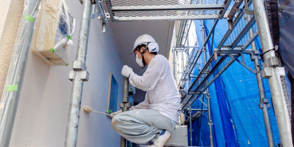  足場上でマスクとヘルメットを着用し、ローラーで外壁を塗装する職人の作業シーン