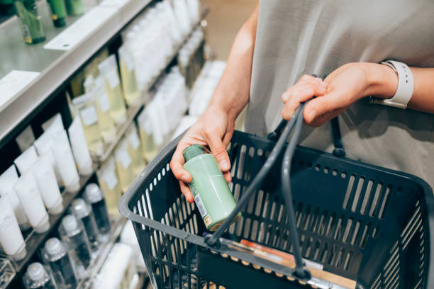 Woman holding a green skincare bottle in a shopping basket while browsing personal care products in a store aisle.