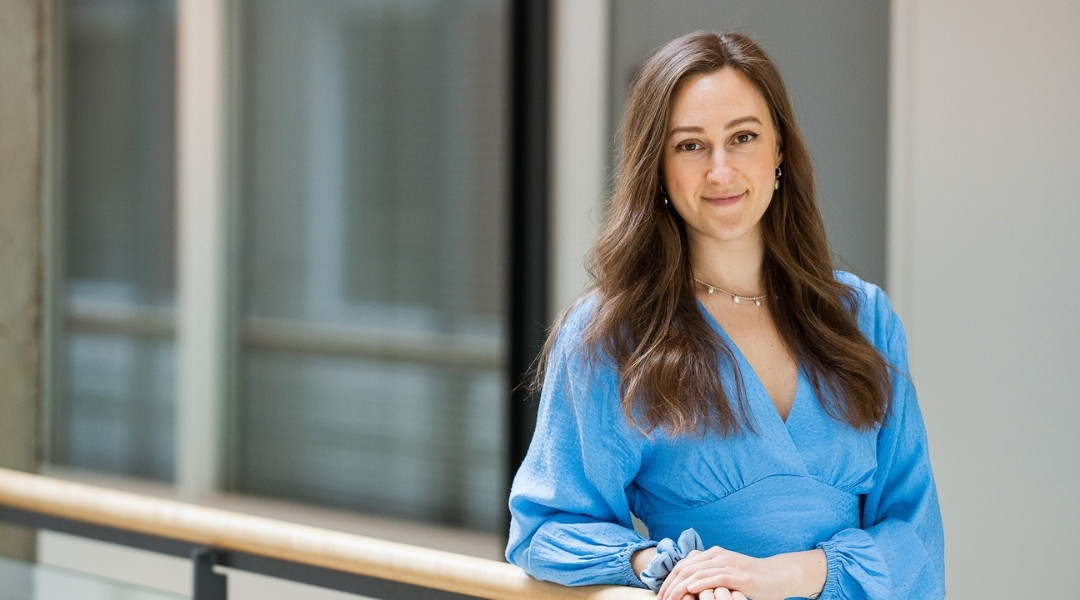 Une nutritionniste à shawinigan souriante portant une blouse bleu clair, debout à l'intérieur près d'une rambarde, avec un éclairage doux et un arrière-plan moderne flou.