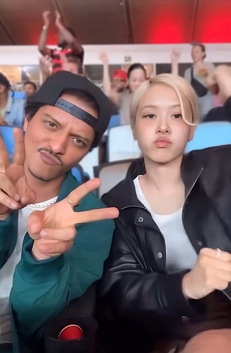 This contains an image of BLACKPINK's Rosé and Bruno Mars sitting next to each other in front of a crowd with one pointing at the camera