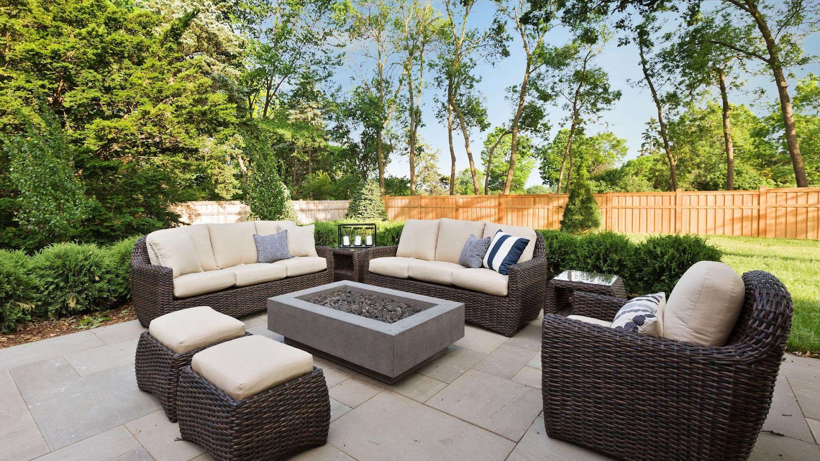 A cozy backyard setup with woven furniture and a central concrete fire pit, surrounded by lush greenery and a wooden fence, showcasing a modern and inviting outdoor living space.