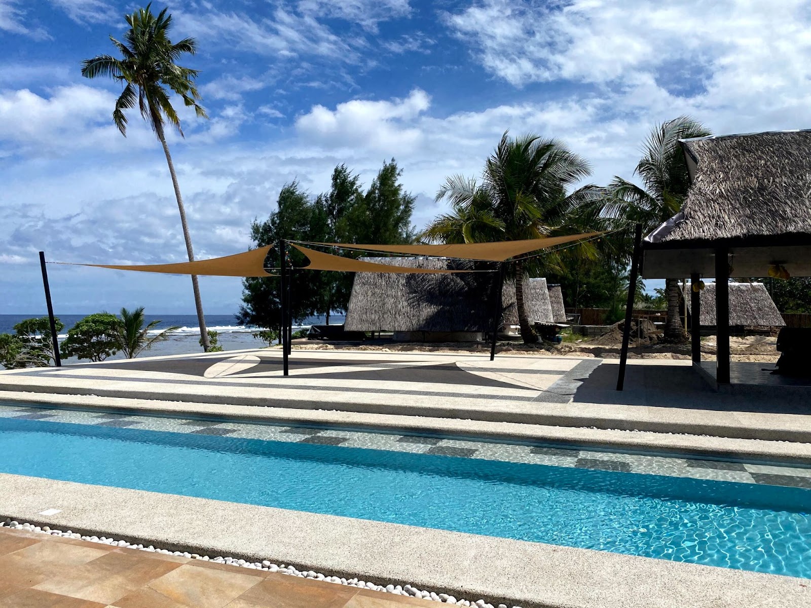 Luxury beachfront poolside with tropical huts and palm trees under a sunny sky.