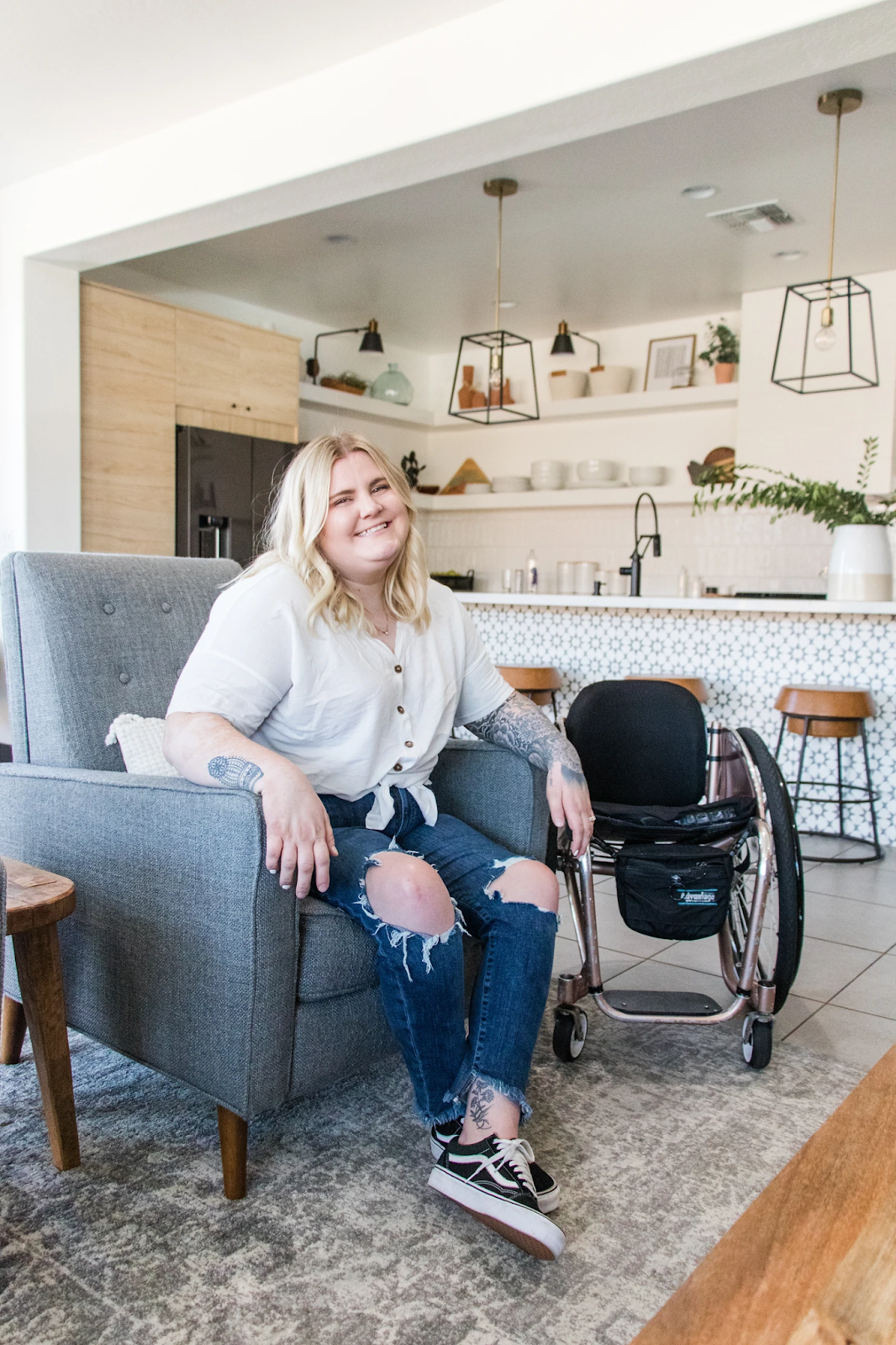 A woman with blonde hair, wearing a white blouse and ripped jeans, is smiling while seated in a gray armchair. Her wheelchair is positioned beside her in a modern, accessible kitchen and living space with open shelving, patterned tile, and midcentury-style lighting.