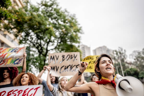 People at a protest holding signs, one reads &quot;My Body, My Choice.&quot; A person in front has a megaphone, wearing casual attire with a scarf