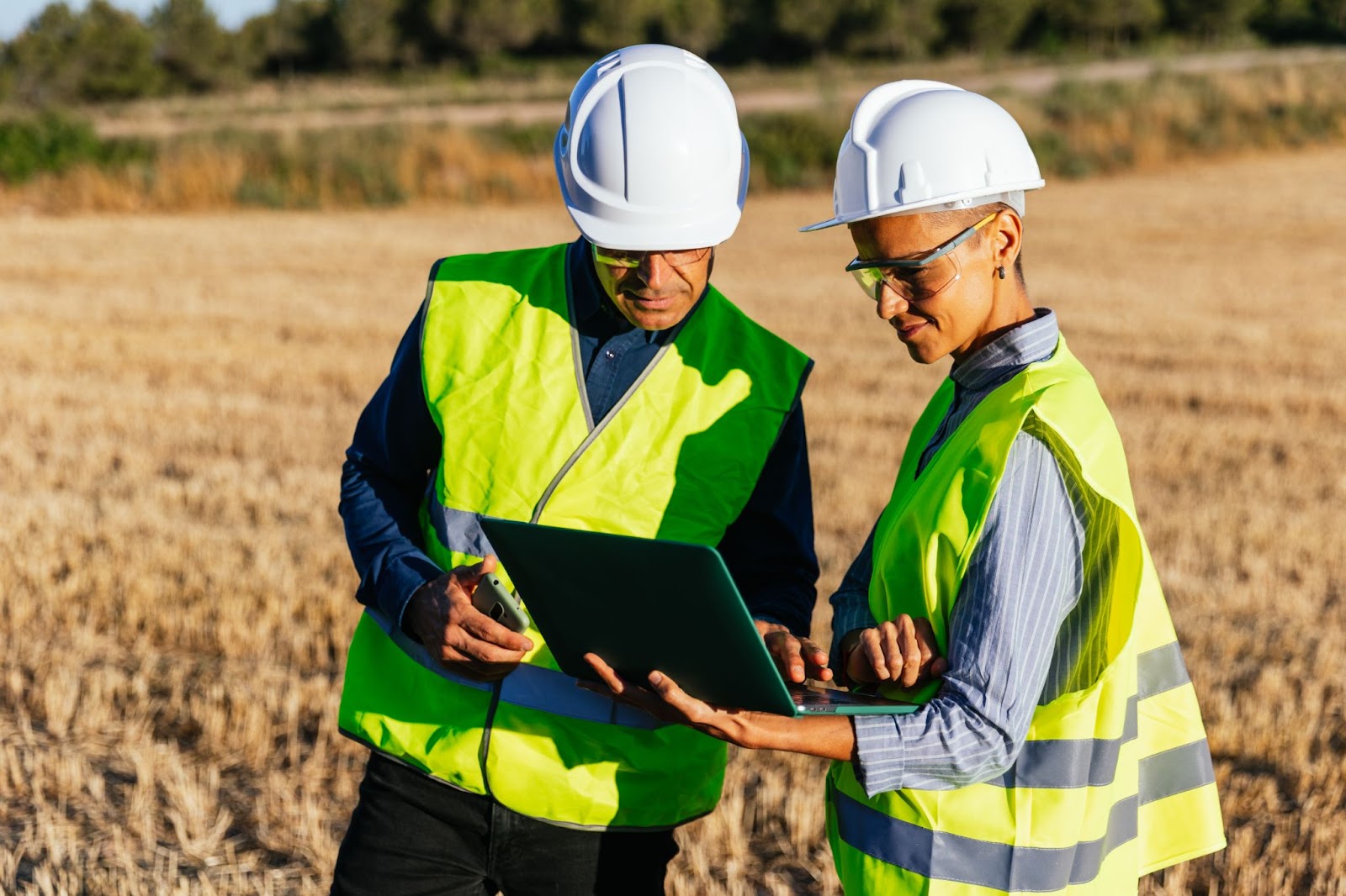 Man and a woman in hard hats and vests using a laptop while in a field. 